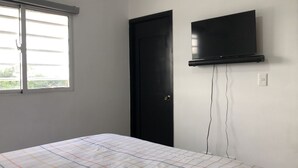 Un dormitorio con cama, una puerta negra, televisor montado en la pared y ventana con persianas.