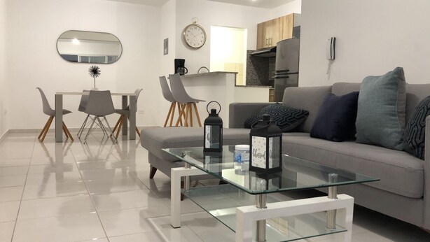 Una sala moderna con una mesa de centro de vidrio, un sofá gris y una zona de comedor con sillas y una mesa pequeña.