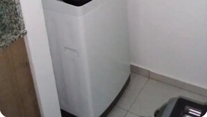 Lavadora blanca compacta en una habitación pequeña con piso de baldosas y una tubería de agua visible.
