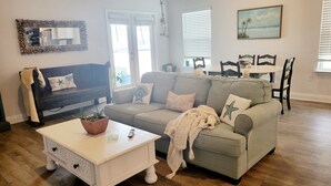 Un salon avec un canapé gris, une table basse blanche ornée d’une plante et un tableau représentant une scène de plage.