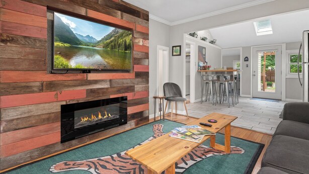 A living room with a fireplace, a television mounted on a wooden wall, a dining area with bar stools, and a view of a mountain lake through a window. - Music Lover’s Getaway - Red Rocks + Denver (Wheat Ridge)