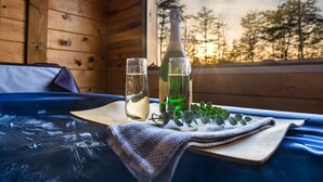 Ein Holzblockhaus-Interieur mit einem Holzbett, einem Fenster mit Blick auf Bäume, einer Flasche mit grüner Flüssigkeit und einem Glas mit klarer Flüssigkeit auf einem Holztisch.