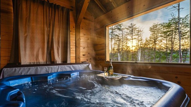 Ein Whirlpool in einer Holzhütte mit Blick durch ein Fenster auf Bäume und einen Sonnenuntergang.