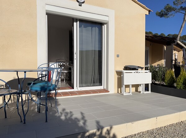 Une terrasse avec une table et des chaises, une porte-fenêtre coulissante et une petite table sur laquelle sont posés divers objets.