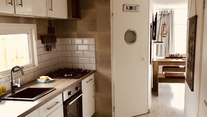 Une cuisine moderne avec des meubles blancs, un plan de travail noir et un four encastré. Il y a une fenêtre avec des stores et une porte portant l’inscription « KOJE ».
