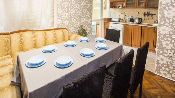 Uma sala de jantar com mesa posta para quatro pessoas, um sofá e uma cozinha ao fundo.