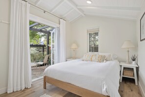 A bedroom with a large bed, two bedside tables, a window, and a view of an outdoor patio.