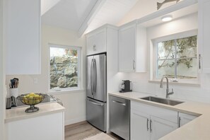 A modern kitchen with white cabinetry, a stainless steel refrigerator, a dishwasher, and a window with a view of greenery.