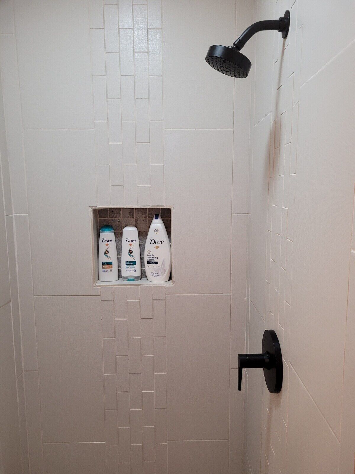 A shower with a black handheld showerhead, a tiled wall, and a shelf with Dove body wash bottles.
