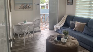 A cozy living room with a blue sofa, a white coffee table, and a patterned ottoman. There is a dining area with a small round table and chairs. A painting hangs on the wall, and a sign above the door reads "Bless this Home with Love & Laughter."