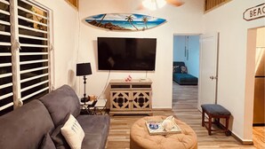 A living room with a sofa, ottoman, TV, and a painting of a beach scene.