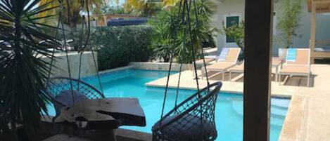 Un espace piscine agrémenté d’une chaise suspendue, de chaises longues et d’un petit jardin avec des plantes tropicales.