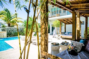 Un espace aménagé au bord de la piscine, avec une pergola en bois, des chaises longues et une table où sont servis des plats et des boissons.