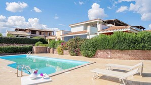 Un'area piscina con una piscina blu cristallina, una poltrona da relax bianca e un tavolo bianco con un vaso e fiori.