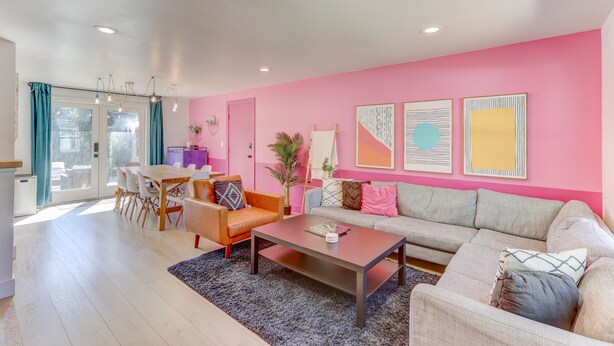 A modern living room with a pink wall, a grey sofa, a wooden dining table, and a coffee table. - Spacious 5-Bedroom Retreat - Hot Tub - Games (Denver)