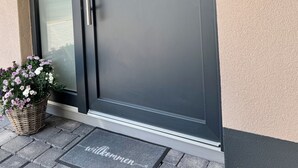 Entrada moderna de un edificio con puerta corredera de vidrio, felpudo y una pequeña lámpara exterior.
