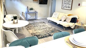 Un salon moderne avec un canapé blanc, des fauteuils turquoise, une petite table ronde et une table basse. Un téléviseur est fixé au mur, et une pancarte indique « MAISON DE PLAGE ».