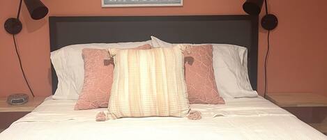 Quarto com cama, duas luminárias de cabeceira, relógio de parede e um letreiro sobre a cama.