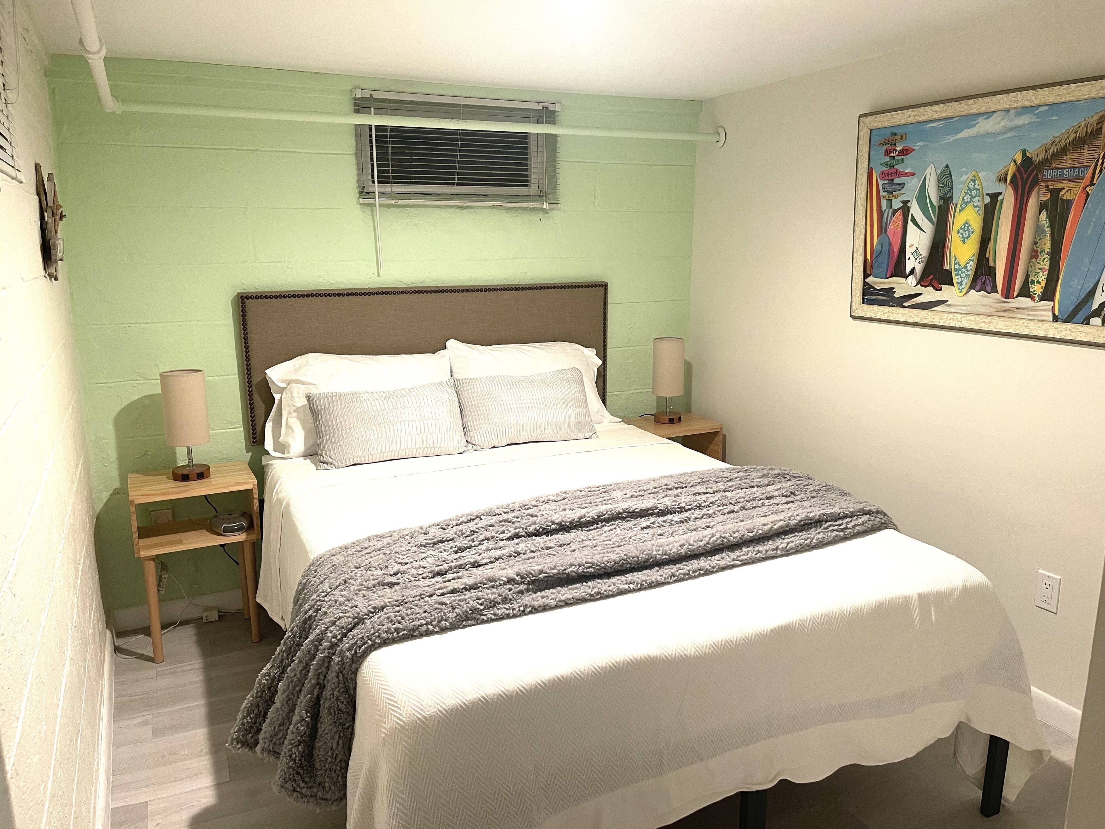 A bedroom with a bed, bedside lamps, a wall-mounted air conditioner, and a surfboard-themed painting.
