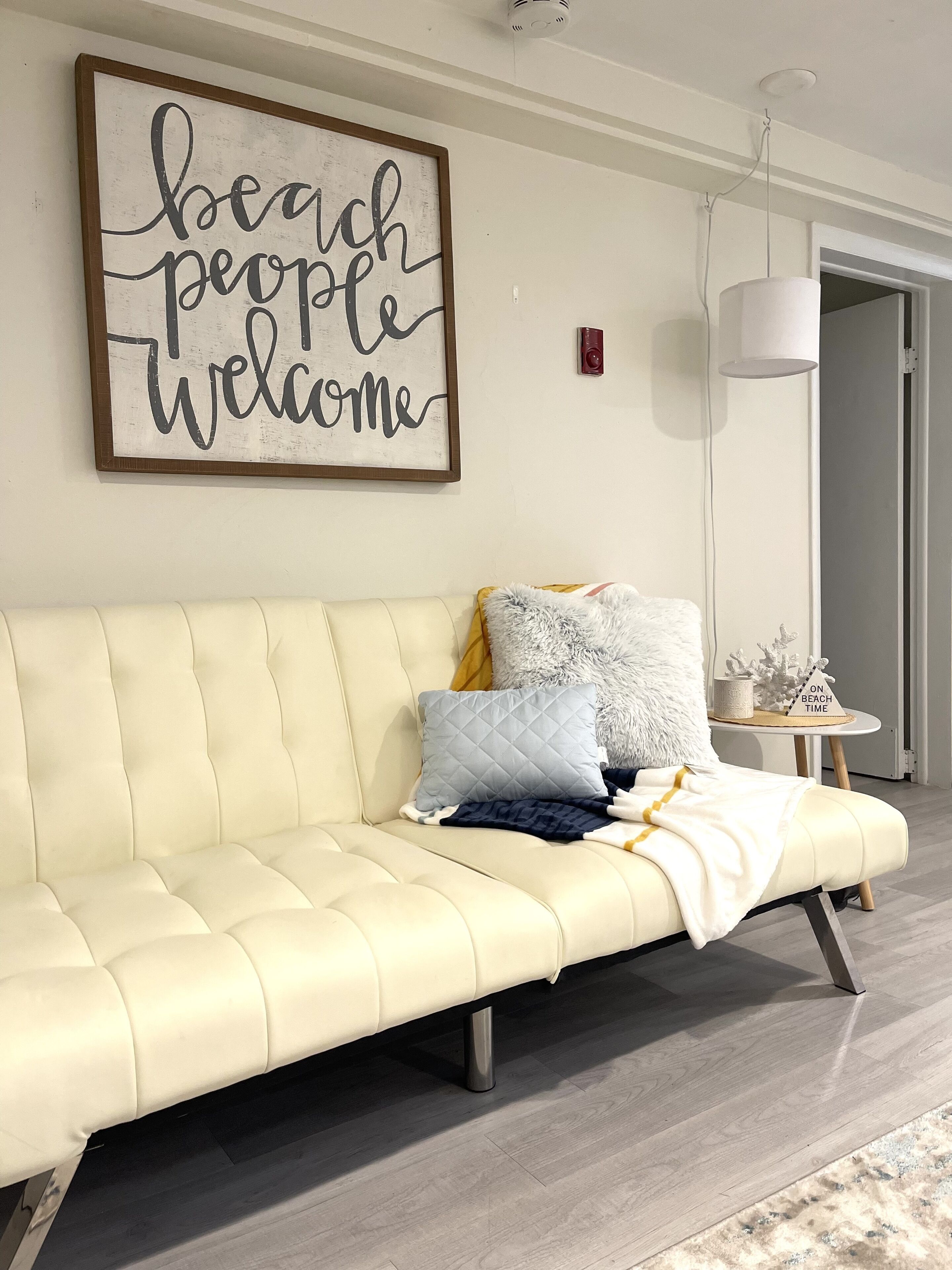 Un sofá color crema con un cojín acolchado y una manta, un letrero enmarcado encima, y una mesita con un objeto decorativo.