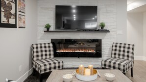 A living room with a fireplace, two checkered armchairs, a TV mounted above the fireplace, and a bowl of popcorn on the table. - 2 Bedroom Bsmt Suite in the Heart of the City (Lethbridge)