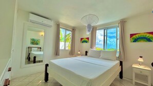 A bedroom with a large bed, a bedside table, a nightstand, a window with curtains, a ceiling fan, and a wall-mounted air conditioner. - Palm House (Grenadines)