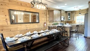 Une salle à manger aux murs lambrissés, avec une longue table dressée pour un repas, un plancher en bois et une cuisine en arrière-plan.