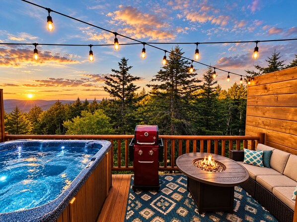 A deck with a hot tub, a barbecue grill, a fire pit, outdoor seating, and string lights against a backdrop of trees and a sunset.