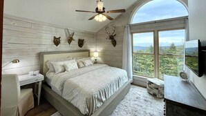 Une chambre à coucher comprenant un lit, un bureau, une chaise, un ventilateur de plafond, une télévision et une grande fenêtre donnant sur des arbres.