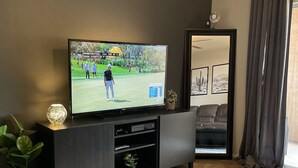 Una sala moderna con una televisión de pantalla plana sobre una consola de madera oscura, sofás de cuero, una planta en maceta y un amplio ventanal con cortinas.