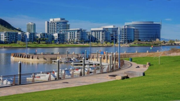 Una zona frente al agua con un puerto deportivo, edificios modernos y un parque arbolado.