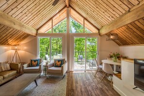 A spacious attic room with a sloped wooden ceiling, large windows, a cozy seating area with a sofa and chairs, a dining table, and a TV. - SLOPESIDE 4 BED,HOT TUB, BEACH, FOUR SEASON, NEAR ALGONQUIN PARK AND ARROWHEAD. (Huntsville)