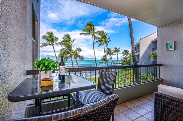 A balcony with a glass table, wicker chairs, and a view of the ocean and palm trees.