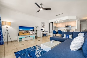 A modern living room with a large flat-screen TV, a ceiling fan, a sofa with decorative pillows, a coffee table, and a blue rug.