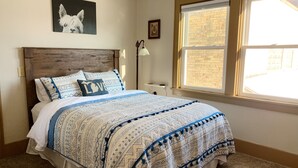 A bedroom with a wooden headboard, a patterned bedspread, a framed picture on the wall, and a window with a view of a brick building. - 2-Bedroom Luxury Loft in the Heart of Downtown Munising (Munising)