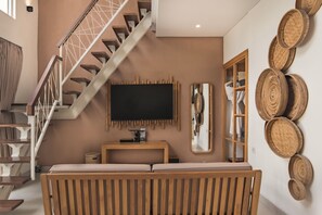 A modern living room with a staircase, a flat-screen TV mounted on the wall, a wooden console table, and a large mirror. - Aeera Villa Canggu by Ini Vie Hospitality (Canggu)