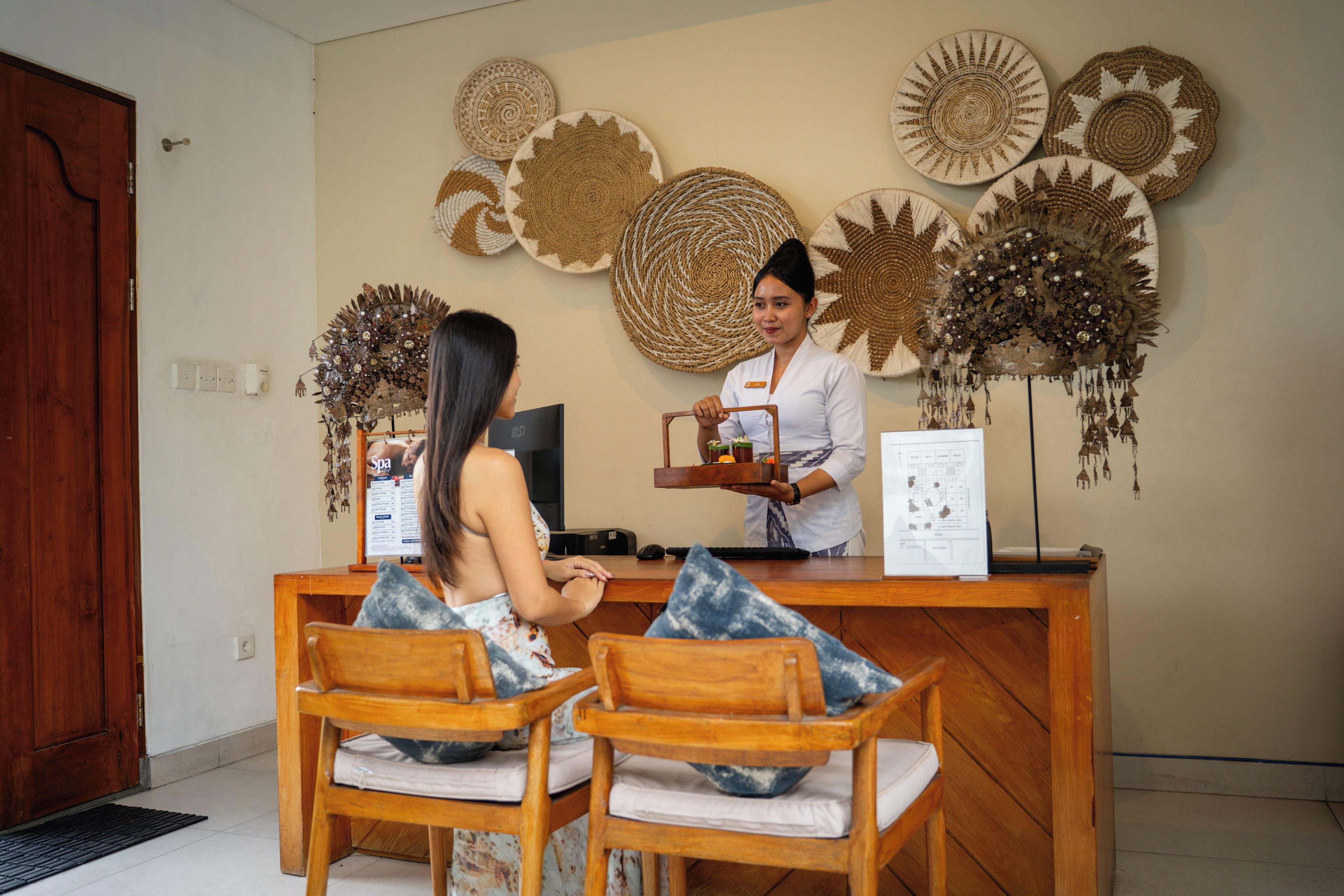 Un mostrador de recepción con detalles de madera, una pared decorada con patrones circulares tejidos y dos sillas de madera con cojines.