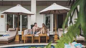 A poolside area with two lounge chairs under umbrellas, a wooden table, and a view of the pool.
