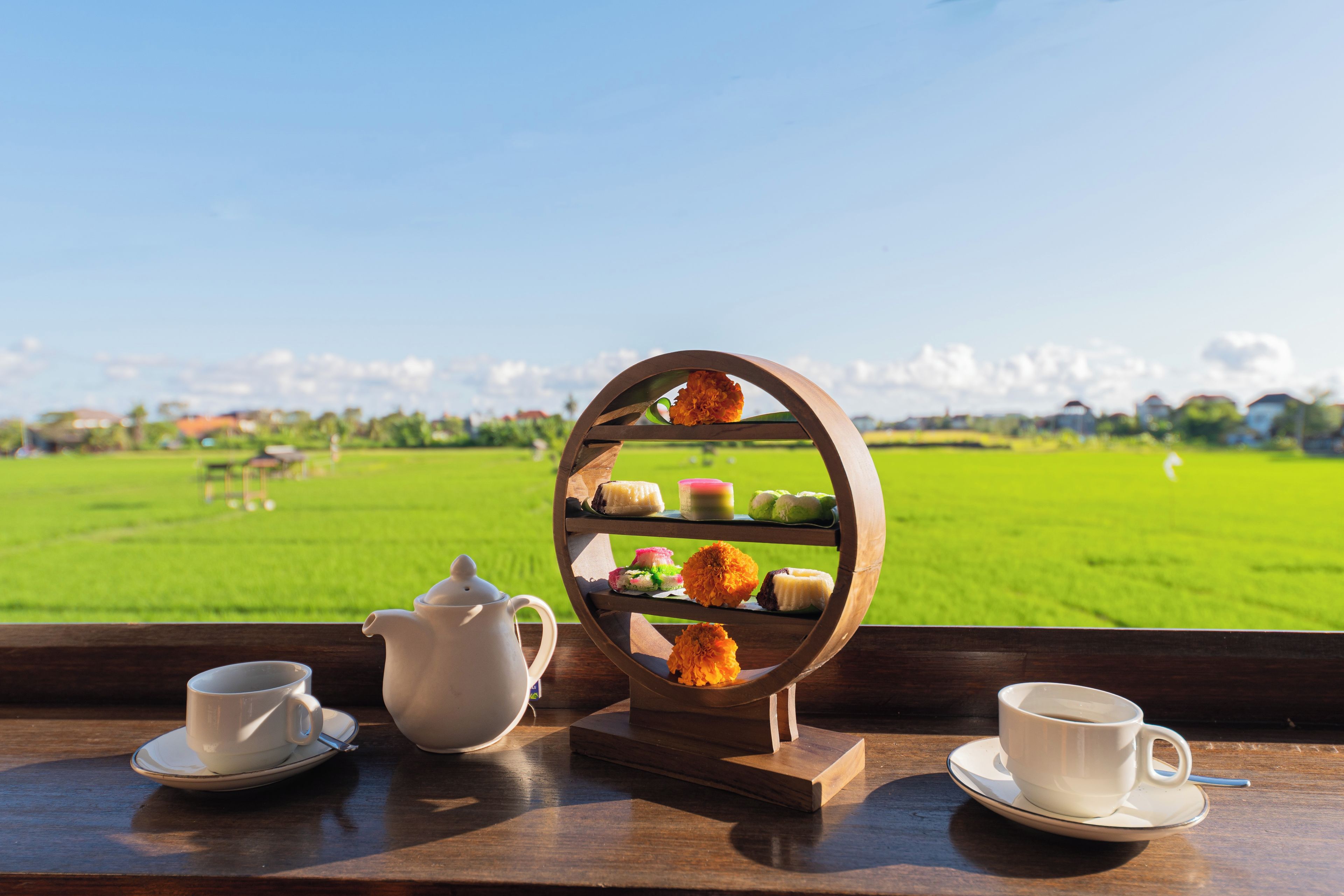 A table with a tiered stand holding various desserts, two white cups, and a white teapot, set against a view of a green field and a clear sky.