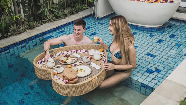 Escena junto a la piscina con un jacuzzi, una mesa de comedor con comida y bebidas, y una persona sentada en la piscina.