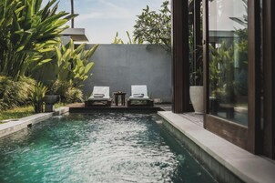 Zona de piscina con dos sillas reclinables y una mesa, rodeada de vegetación y un edificio moderno.