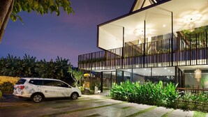 Uma casa moderna de dois andares com fachada de vidro, uma varanda e um carro estacionado na frente.