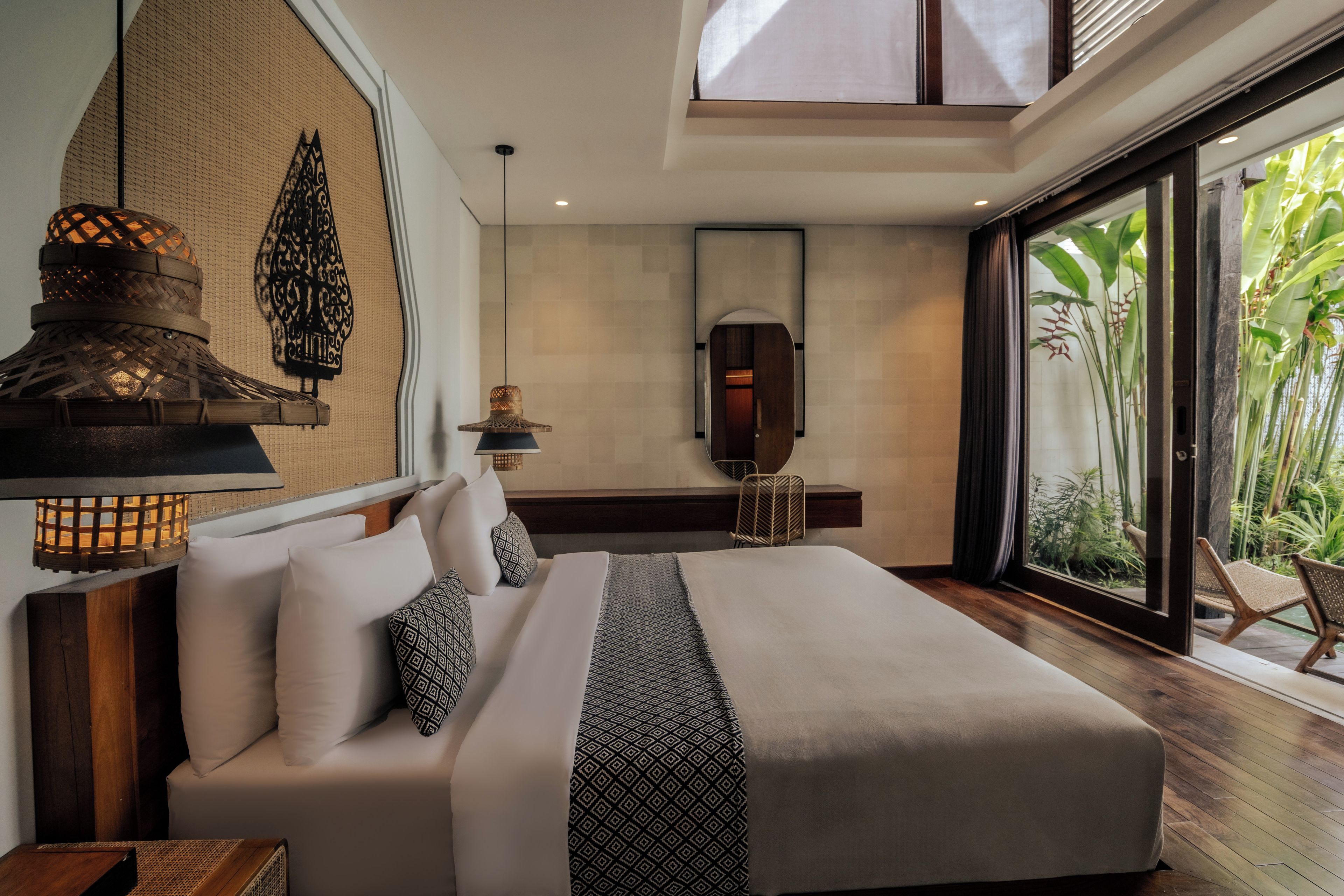 A modern bedroom with a large bed, a hanging light fixture, a mirror, and a view of greenery outside.