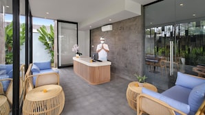 A modern reception area with a wooden reception desk, wicker furniture, and large windows offering a view of the outdoors.
