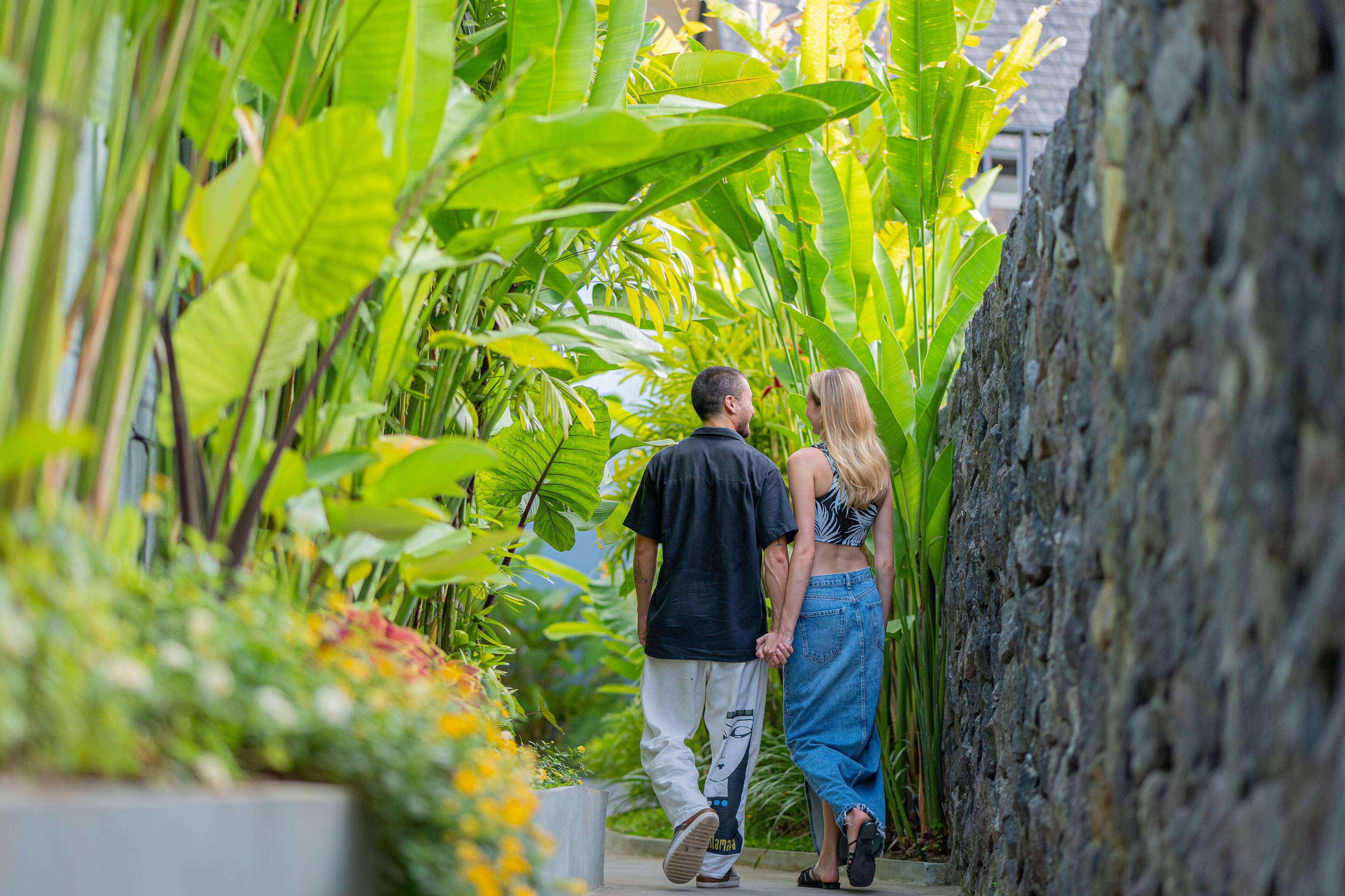 Una pareja caminando por un sendero estrecho rodeado de abundante vegetación y un muro de piedra.