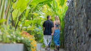A couple walking through a narrow pathway surrounded by lush greenery and a stone wall.
