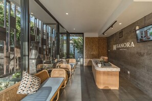 Un área de recepción moderna con mostrador, zona de estar con mobiliario de mimbre y amplios ventanales con vistas a la vegetación.