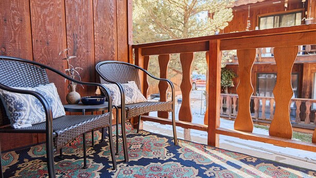 Eine hölzerne Veranda mit zwei Korbstühlen, einem gemusterten Teppich und Blick auf ein Gebäude mit einem Balkon.