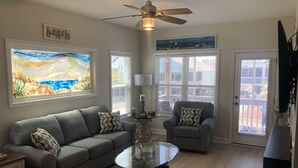 A living room with a gray sofa, a glass coffee table, and a gray armchair. There are two windows with blinds, a ceiling fan, and a painting on the wall.