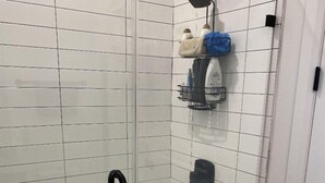 A shower area with white tiled walls, a glass door, and a black handle. There are shelves with toiletries and a towel rack. - TOP FLOOR DOWNTOWN CONDO (Columbus)